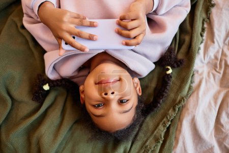 Foto de Retrato de vista superior de una chica negra mirando la cámara boca abajo mientras está acostada en la cama con un smartphone - Imagen libre de derechos