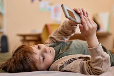 Téléchargez les photos : Portrait de garçon mignon tenant smartphone tout en étant couché sur le lit et souriant, gen Alpha - en image libre de droit
