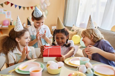Téléchargez les photos : Groupe diversifié d'enfants heureux à la fête d'anniversaire avec des cadeaux d'ouverture de petite fille noire - en image libre de droit