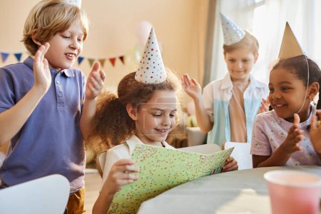 Téléchargez les photos : Groupe diversifié d'enfants heureux à la fête d'anniversaire avec des cadeaux d'ouverture de fille mignonne - en image libre de droit