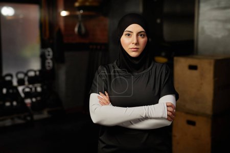 Photo for Young Muslim female fitness instructor in hijab looking at camera while standing in gym against sports equipment and crossing arms by chest - Royalty Free Image