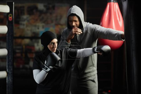 Téléchargez les photos : Jeune femme musulmane en vêtements de sport, hijab et gants de boxe frappant sac de boxe en cuir noir tout en se tenant à côté de l'instructeur masculin - en image libre de droit