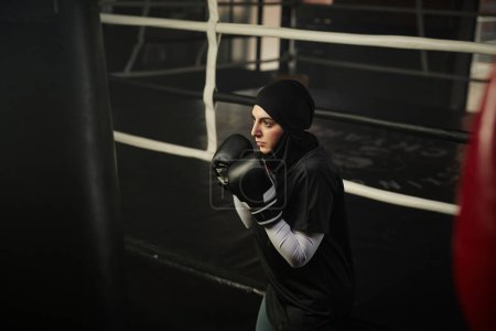 Photo for Young serious Muslim female boxer in activewear and hijab looking at punching bag during training and concentrating on further hit - Royalty Free Image