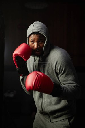Téléchargez les photos : Jeune Afro-Américain en gants de boxe rouges regardant sac de boxe tout en se tenant devant la caméra dans la salle de gym et ayant l'entraînement - en image libre de droit