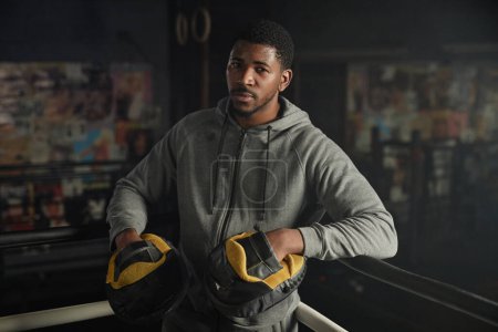 Photo for Young African American male boxing trainer in hoodie and pads standing on rink and looking at camera while having break between trainings - Royalty Free Image