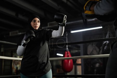Téléchargez les photos : Jeune femme sérieuse en hijab, vêtements de sport et gants de boxe attaquant rivale tout en se tenant devant son entraîneur avec des coussinets sur les mains - en image libre de droit