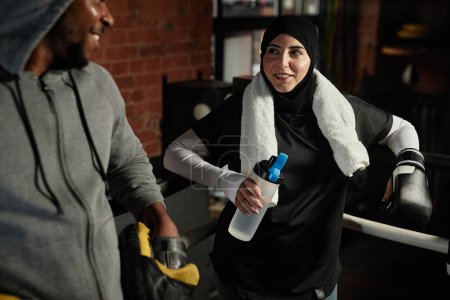 Foto de Feliz joven musulmana con botella de agua mirando al entrenador de boxeo masculino afroamericano en ropa deportiva gris durante el descanso - Imagen libre de derechos