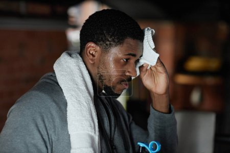 Téléchargez les photos : Jeune boxeur afro-américain fatigué essuyant son front mouillé avec une serviette douce blanche à la pause après un entraînement dur dans une salle de gym ou un club de sport - en image libre de droit