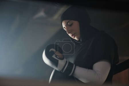 Foto de Joven boxeadora musulmana en hijab poniéndose guantes de boxeo antes de pelear con su rival o entrenando mientras está de pie frente a la cámara en el gimnasio oscuro - Imagen libre de derechos