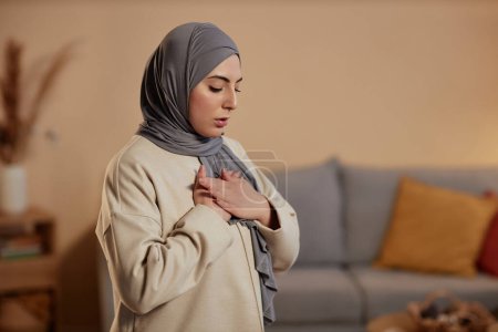 Foto de Joven mujer islámica en hijab gris y sudadera beige expresando gratitud a su Dios durante la oración mientras mantiene sus manos en el pecho - Imagen libre de derechos