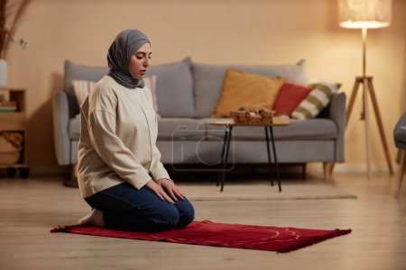 Foto de Joven musulmana en hiyab manteniendo los ojos cerrados mientras se arrodilla sobre una pequeña alfombra roja en la sala de estar y rezando a Allah - Imagen libre de derechos