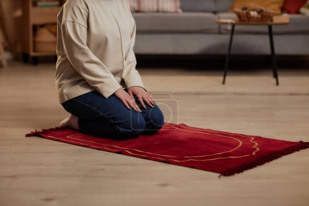 Schnappschuss einer jungen islamischen Frau in Freizeitkleidung, die auf einem kleinen roten Teppich im Wohnzimmer kniet, während sie morgens Gebete zu Gott spricht