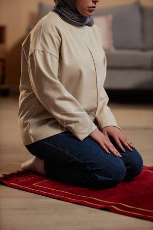 Cropped shot of young woman in beige pullover and blue jeans saying prayers to Allah while standing on her knees on small red rug