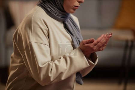 Nahaufnahme einer jungen Muslimin in grauem Hijab und beigem Pullover, die im Wohnzimmer betet, während sie offene Handflächen vor sich hält