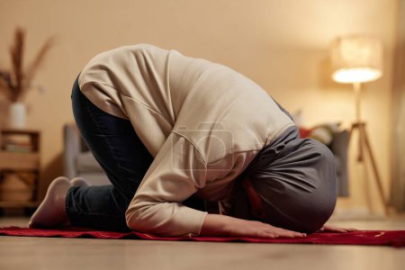 Foto de Vista lateral de la joven musulmana en el hiyab tradicional inclinándose hacia adelante mientras se arrodilla en una pequeña alfombra roja en el ambiente hogareño y rezando - Imagen libre de derechos