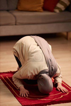 Téléchargez les photos : Jeune femme musulmane en pull beige et hijab gris priant sur un petit tapis rouge tout en s'agenouillant sur le sol à la maison et en se penchant vers l'avant - en image libre de droit
