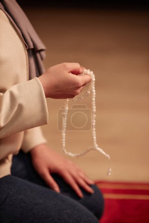 Nahaufnahme der Hand einer jungen islamischen Frau mit weißem Rosenkranz, die zu Hause vor der Kamera kniet und schweigend betet