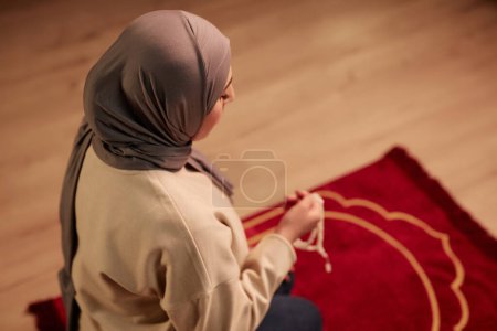 Enfócate en la joven musulmana en hijab gris y jersey beige de rodillas sobre una alfombra roja en casa y practicando la oración silenciosa
