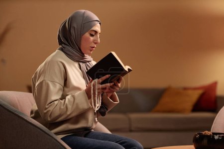 Junge ernsthafte islamische Frau in Hijab und Freizeitbekleidung sitzt im Sessel vor der Kamera und liest Quran, während sie den Tag zu Hause verbringt