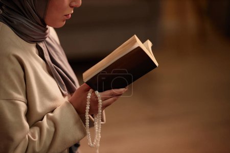 Primer plano de la joven mujer islámica en jersey beige y pañuelo gris en la cabeza sosteniendo quran abierto delante de sí misma mientras lee versos