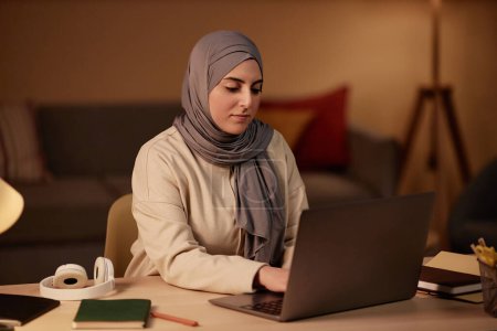 Junge muslimische Geschäftsfrau in Hijab und Freizeitbekleidung tippt am Arbeitsplatz im Home Office und beim Netzwerken auf der Laptop-Tastatur