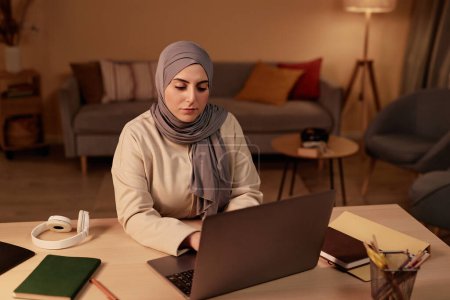 Junge seriöse muslimische Geschäftsfrau sitzt am Arbeitsplatz im Wohnzimmer und schaut auf den Laptop-Bildschirm, während sie durch Online-Daten scrollt