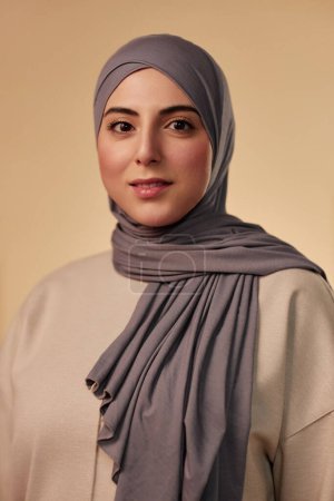 Young beautiful Muslim woman in grey headscarf and beige pullover looking at camera while standing in isolation against pastel background
