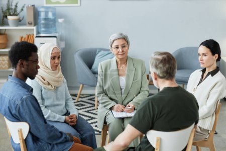 Confident mature female counselor consulting people with psychological problems sitting around her and listening to one of patients at session