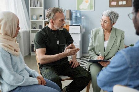 Mature bearded man in casualwear describing his problem to psychologist while sitting among other patients during communication at session