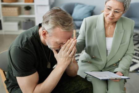 mature man with post traumatic syndrome keeping his hands put together close to face while sitting next to counselor supporting him at session