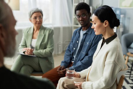Side view of stressed mature brunette woman sharing her problems with support group consisting of psychologist and other patients