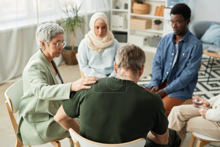 Photo for Rear view of mature stressed man with post traumatic syndrome sitting among intercultural people and psychologist comforting him - Royalty Free Image