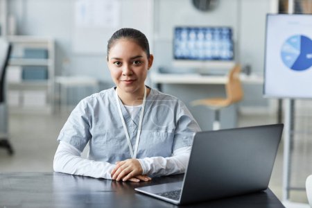 Joven mujer bonita médico o asistente mirando a la cámara mientras está sentado frente a la computadora portátil por el lugar de trabajo y consultar a los pacientes en línea