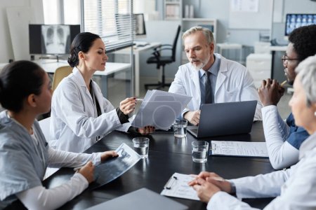 Photo for Young female clinician showing medical documents to experienced male doctor sitting by table in front of group of intercultural colleagues - Royalty Free Image