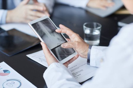 Hände eines reifen männlichen Radiologen blättern durch Röntgenbilder auf dem Tablet-Bildschirm, während er bei einem Meeting mit seinen Kollegen am Tisch sitzt
