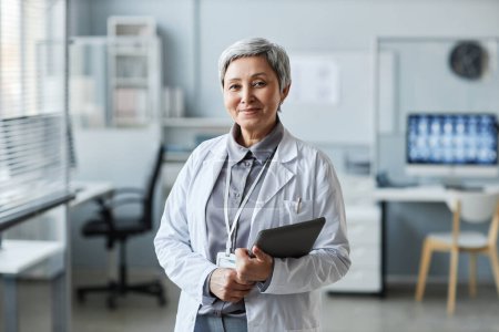 Mujer madura feliz médico con la tableta de pie delante de la cámara en el consultorio médico y mirándolo contra los lugares de trabajo de los colegas