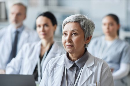 Mature Asian female doctor in lab coat looking at speaker making report opon medical subject while sitting among colleagues at conference