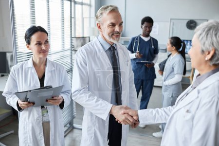 Smiling experienced hospital worker shaking hand of mature female colleague while congratulating her on successful report at seminar