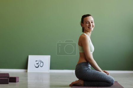 Téléchargez les photos : Vue latérale de la forme jeune femme faisant du yoga à l'intérieur et souriant à la caméra heureusement avec l'espace de copie - en image libre de droit