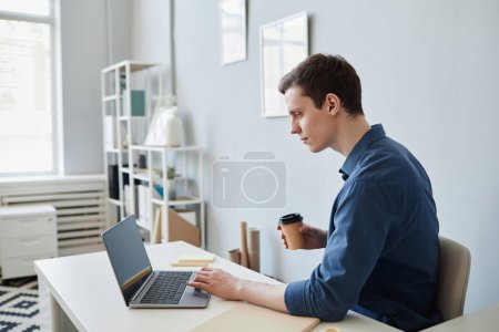 Téléchargez les photos : Portrait latéral d'un jeune homme utilisant un ordinateur au bureau et buvant du café - en image libre de droit