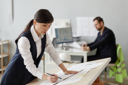 Photo for Side view portrait of young woman as engineer drawing blueprints and plans at table in office, copy space - Royalty Free Image