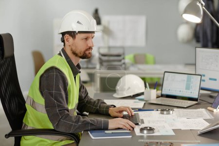Téléchargez les photos : Portrait latéral d'un ingénieur de construction barbu portant un casque de sécurité au travail au bureau et utilisant un ordinateur - en image libre de droit