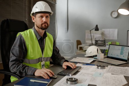 Téléchargez les photos : Portrait d'un ingénieur en mécanique barbu portant un casque de sécurité lorsqu'il utilise un ordinateur au travail au bureau et ayant l'air concentré, espace de copie - en image libre de droit
