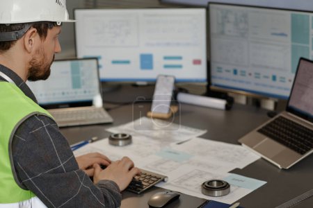 Photo for Side view of male engineer wearing hardhat at workplace in office and using computer designing blueprints, copy space - Royalty Free Image