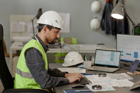 Photo for Side view portrait of male engineer wearing hardhat at workplace in office and using multiple computer devices for calculation and design, copy space - Royalty Free Image