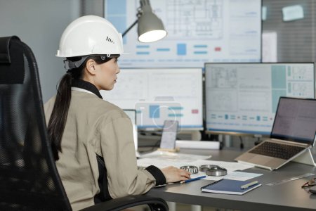 Foto de Retrato de vista lateral de una mujer ingeniera que usa casco en el lugar de trabajo en la oficina y usa computadora con planos en la pantalla, espacio para copiar - Imagen libre de derechos