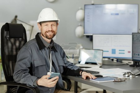 Photo for Portrait of bearded engineer wearing hardhat at workplace in office and looking at camera smiling, copy space - Royalty Free Image