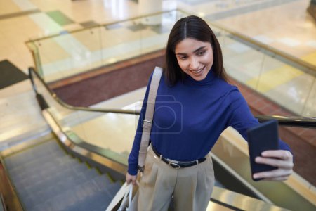 Téléchargez les photos : Portrait en grand angle d'une jeune femme souriante prenant des photos selfie ou filmant une vidéo debout sur un escalier roulant dans un centre commercial et tenant des sacs espace de copie - en image libre de droit