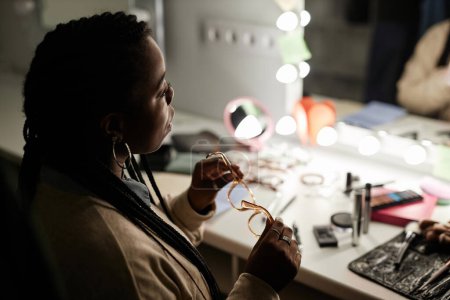 Foto de Retrato de vista lateral de una joven negra sentada en la mesa de tocador en el vestidor y mirando el espejo mientras se prepara para el espacio de copia de rendimiento - Imagen libre de derechos