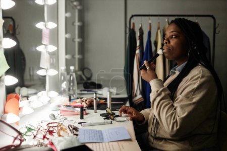 Téléchargez les photos : Vue de côté portrait de la femme afro-américaine faisant maquillage par miroir dans le vestiaire se préparer pour l'espace de copie de performance - en image libre de droit